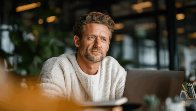 Een man van rond de veertig kijkt nadenkend voor zich uit terwijl hij achter zijn laptop zit in een modern kantoor met veel licht en groen. De foto straalt rust, vertrouwen en focus uit – passend bij het thema investeren in vastgoed.